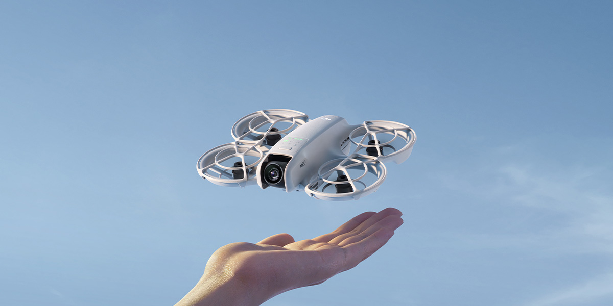 Dron DJI Neo unosi się nad dłonią na tle błękitnego nieba