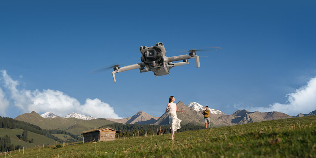 Dron w locie nad łąką w górach, za nim mężczyzna i kobieta